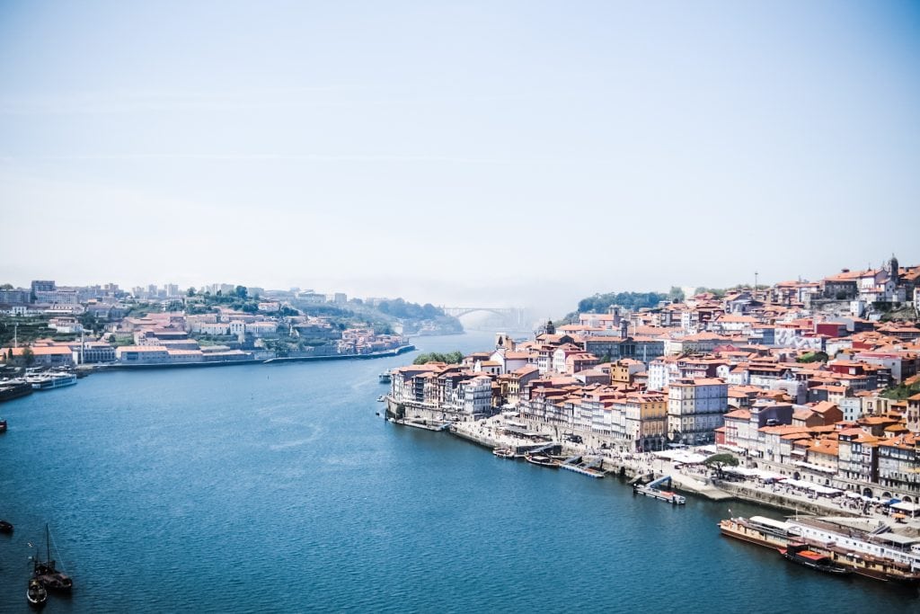 Aerial view of the Douro River from the Dom Luis Bridge flowing between two urban areas with rows of colorful buildings, boats docked along the waterfront.