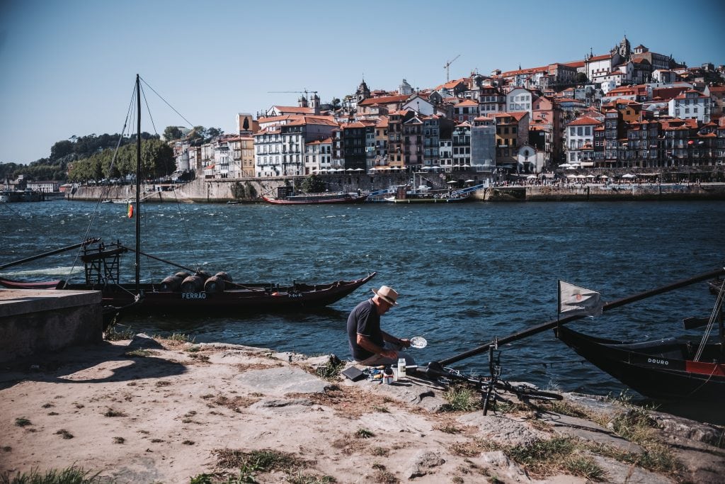 A person sits near the water with boats docked nearby, overlooking the riverside town of Porto with many buildings and a hill in the background along the Douro River.