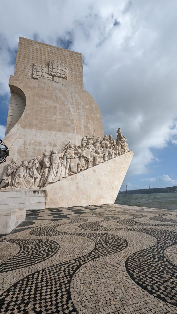 A monumental stone structure featuring detailed sculptures of historic figures, reminiscent of Lisbon's Monument of Discoveries, set against a cloudy sky with a patterned tiled plaza in the foreground and the Tagus river in the background.