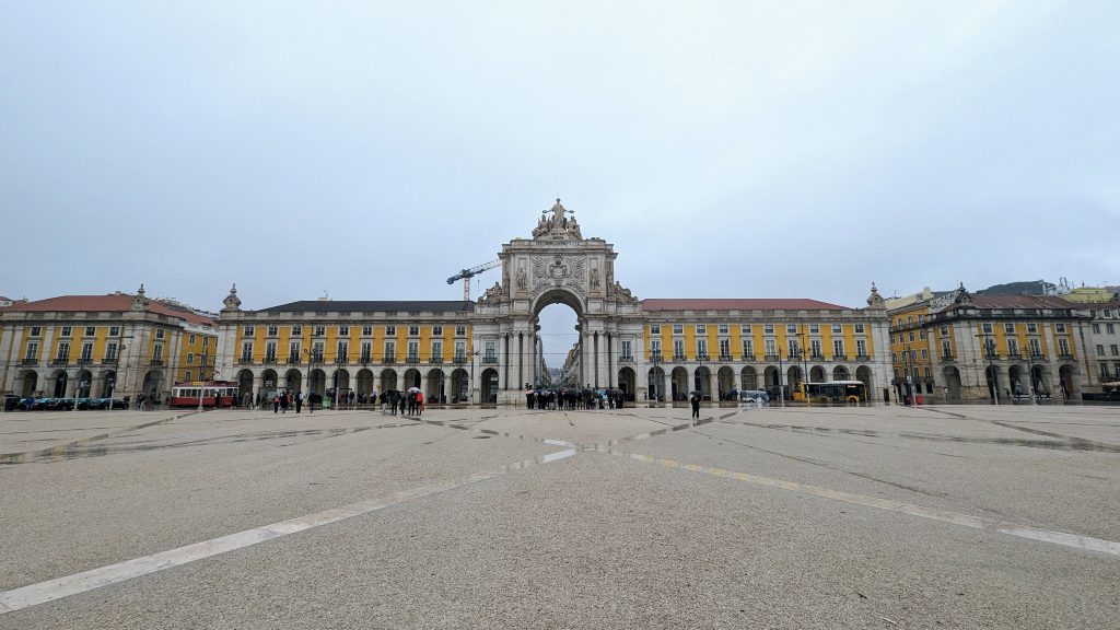 A wide, paved plaza with a large, ornate archway in the center, flanked by symmetrical yellow buildings with arched windows and columns. The sky is overcast. This picturesque setting can be found at Lisbon's iconic Praça do Comércio.