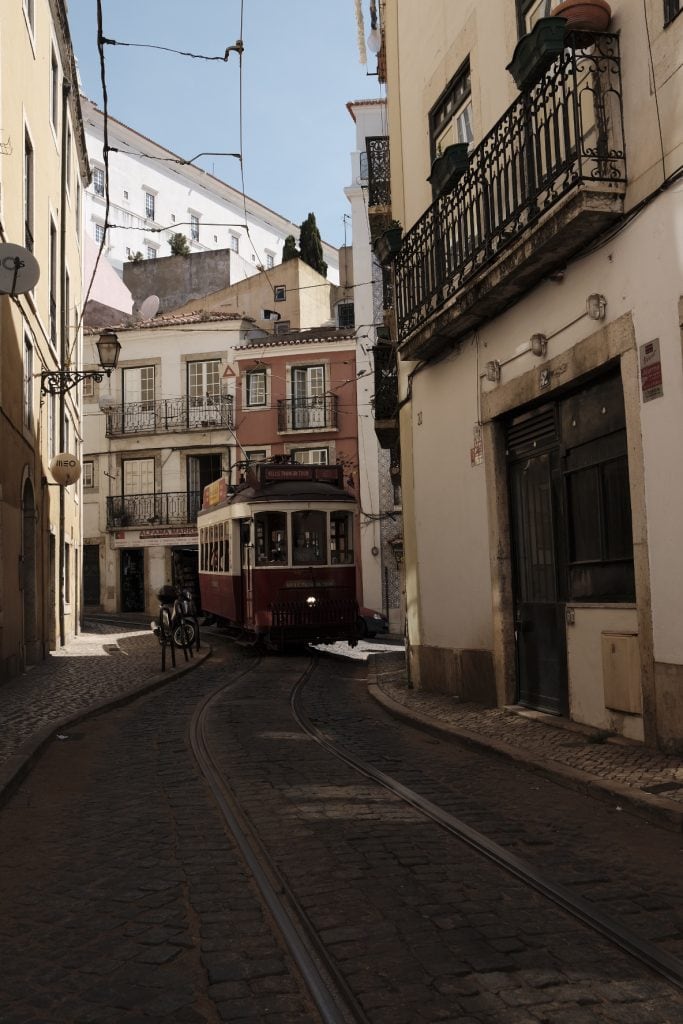 A red tram passes through a narrow, cobblestone street surrounded by historic buildings with balconies in the picturesque city of Lisbon.