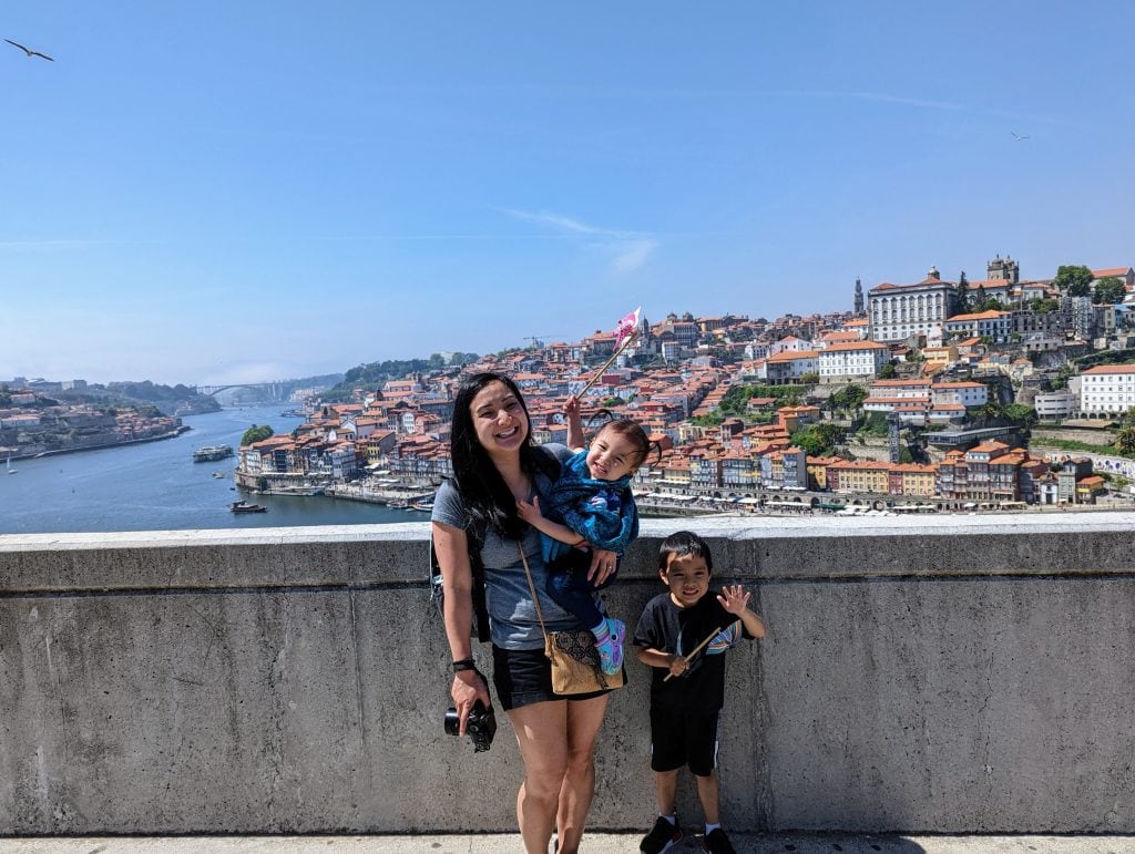 A woman with two children stands on a concrete promenade with a scenic view of the river and Porto's terracotta rooftops in the background. The children hold windmills.