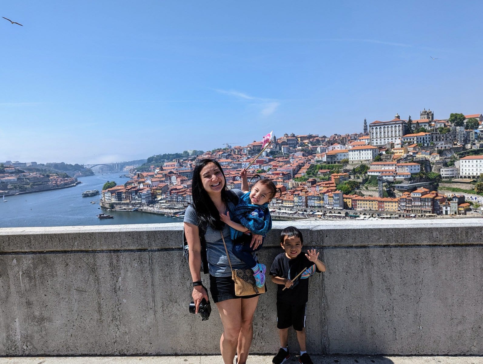A woman with two children stands on a concrete promenade with a scenic view of the river and Porto's terracotta rooftops in the background. The children hold windmills.