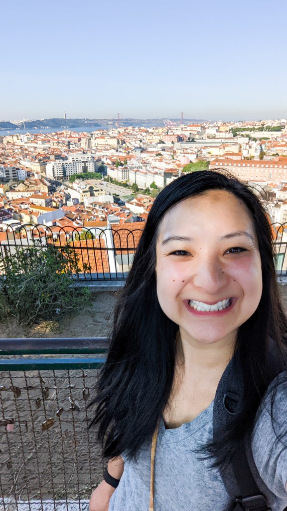 A person with long black hair smiles widely while standing in front of a panoramic view of Lisbon, featuring red-roofed buildings and a distant bridge in the background.