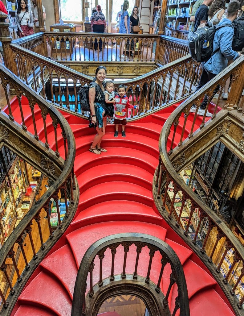 A woman and two children stand on a split staircase with bright red steps in the bookstore Livraria Lello in Porto. People are browsing books in the background.