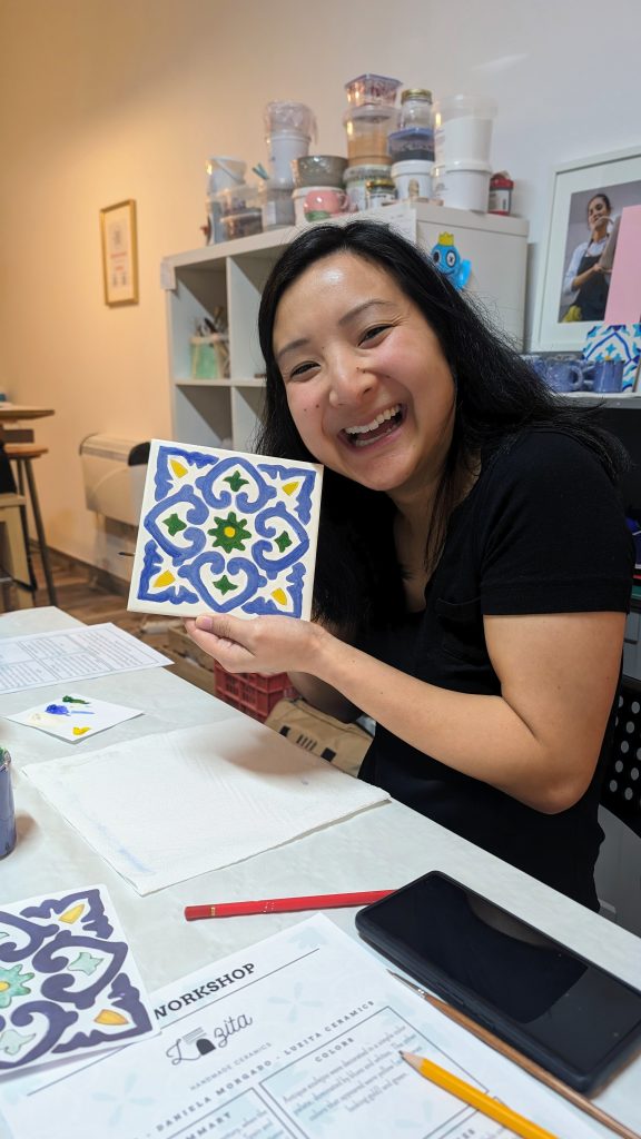 A person smiles while holding a hand-painted tile with a colorful geometric design during a workshop. The table has art supplies and a workshop guide.