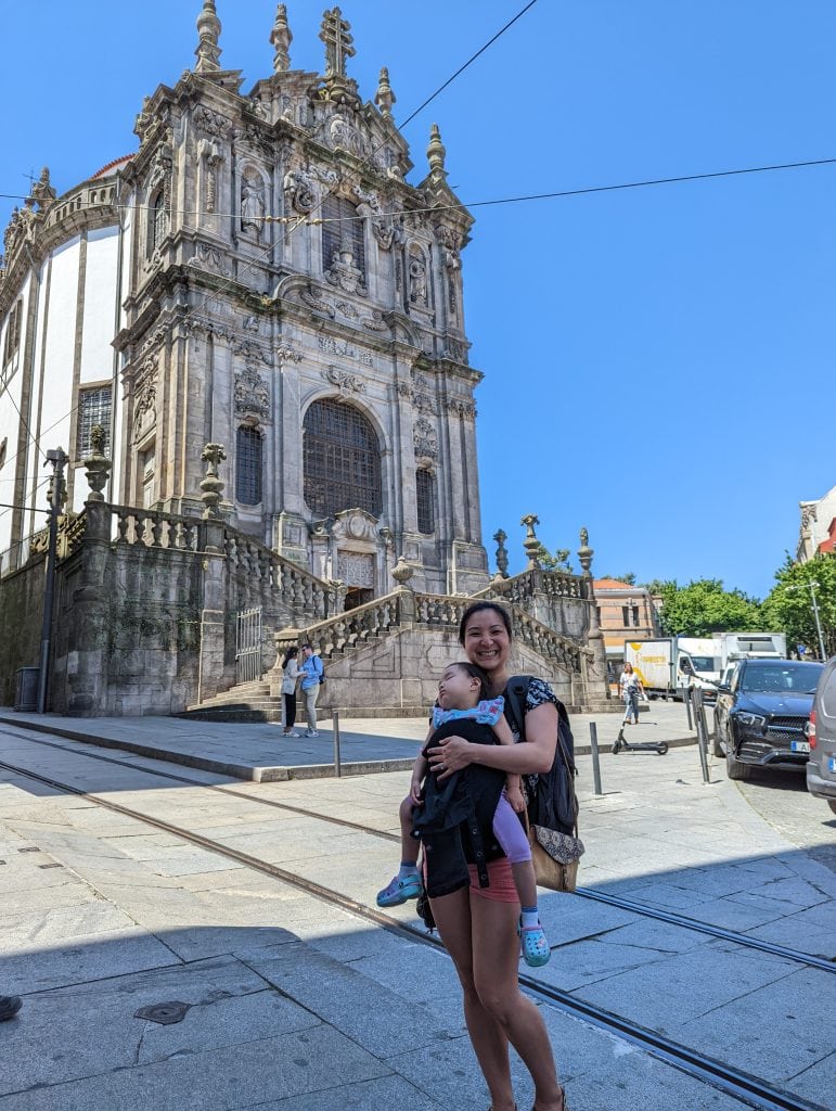 A woman holding a baby stands in front of an ornate historical church, with stairs leading up to its entrance on a sunny day.