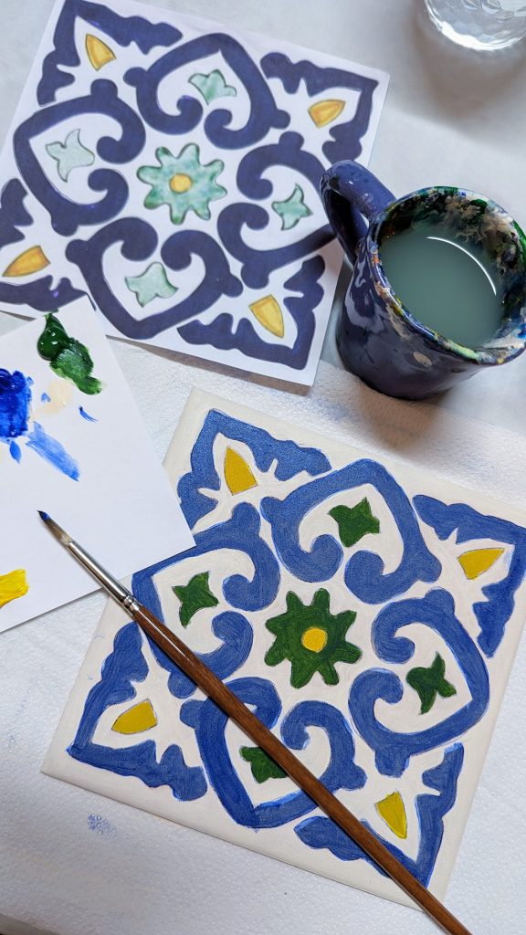 An intricately painted tile are displayed alongside a paintbrush, a small palette with mixed colors, and a cup of water used for cleaning the brush.