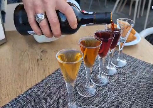 A hand is pouring dark port wine from a bottle into a row of five small glasses on a wooden table, with food items visible in the background.