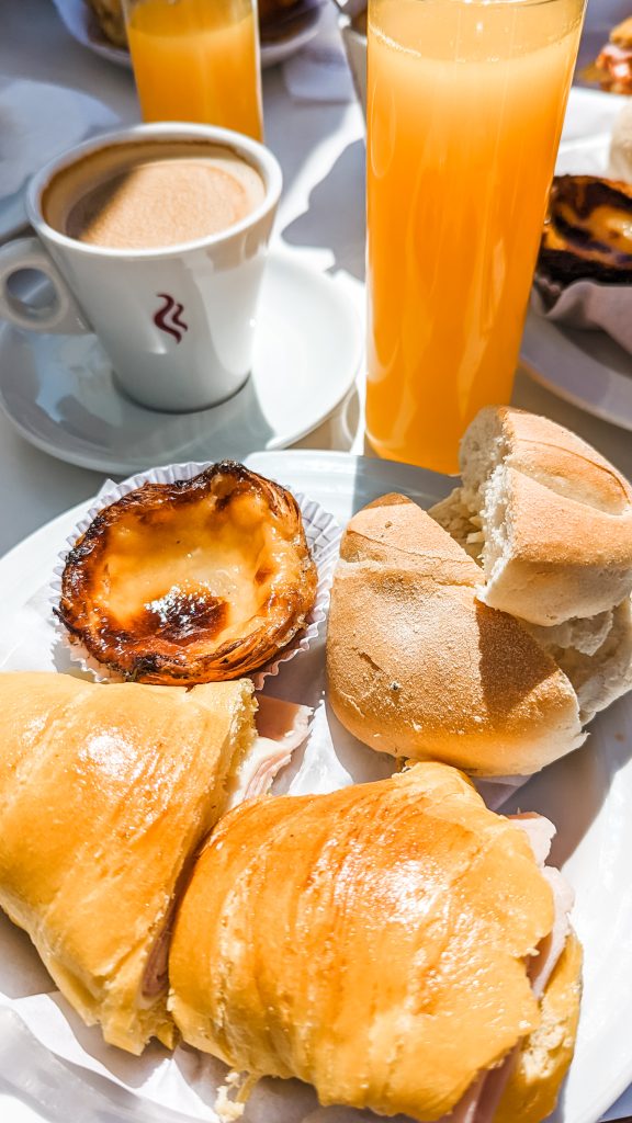 A breakfast spread includes a cup of coffee, a glass of orange juice, a pastel de nata custard tart, a croissant sandwich, and a bread roll on a white plate.