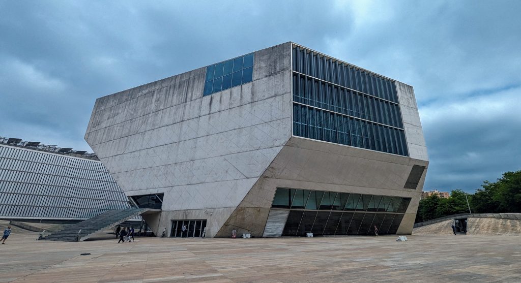 A modern, angular building with large glass windows and concrete walls under a cloudy sky. People are walking near the entrance.