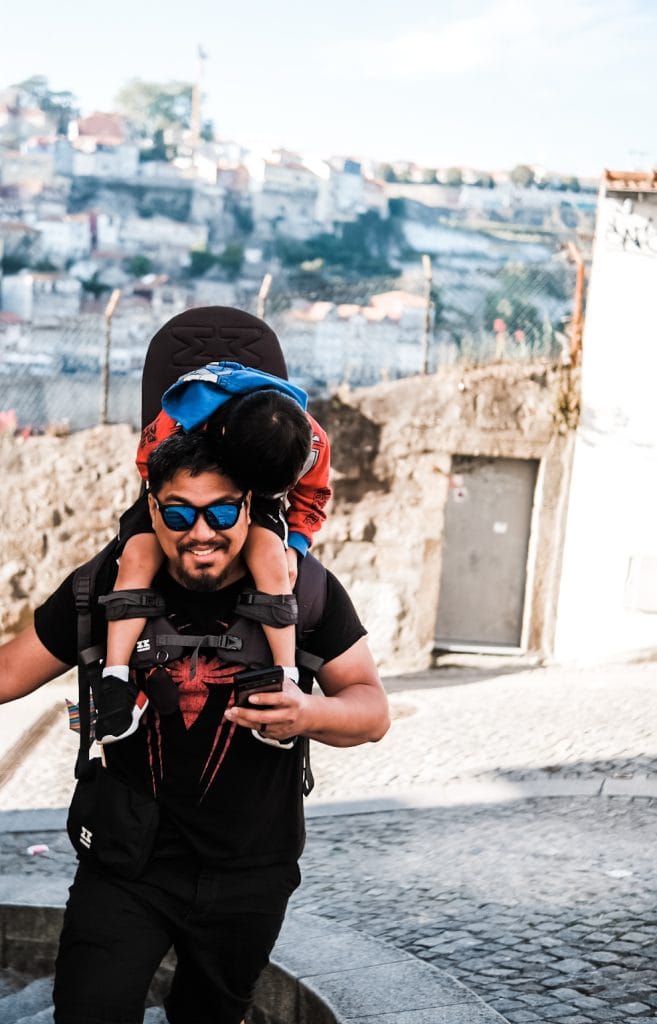 A man wearing sunglasses carries a young child on his back while walking up a cobblestone street. The background features a distant view of buildings and a metal fence.