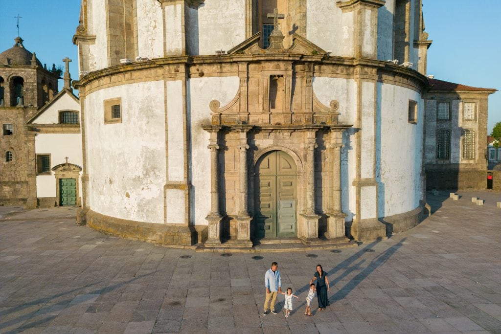 A family of four stands in front of a large, circular stone building with ornate architecture, including an arched doorway and columns. The building is set in a spacious, paved courtyard.