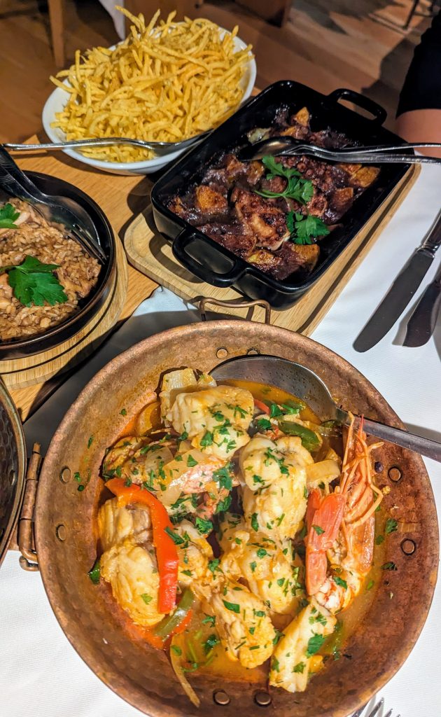 A table set with a plate of seasoned fries, a dish of cooked octopus topped with herbs, and a copper pan filled with seafood and vegetables garnished with parsley.