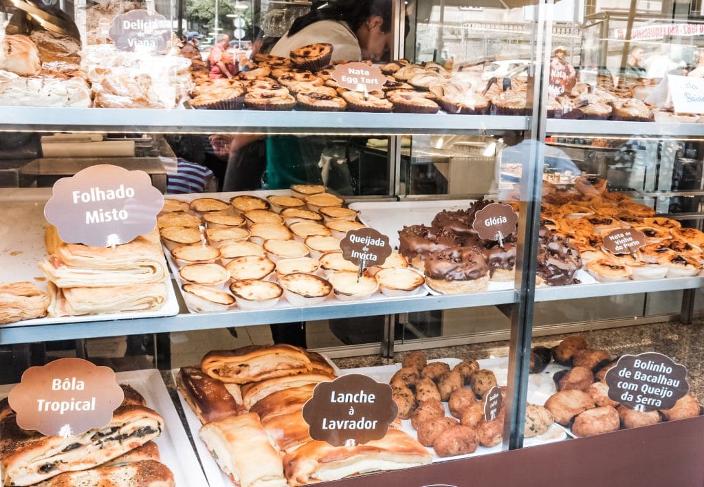 A bakery display case filled with a variety of pastries including folhado misto, bola tropical, and lanche à lavador, all labeled. A person in the background is arranging items.