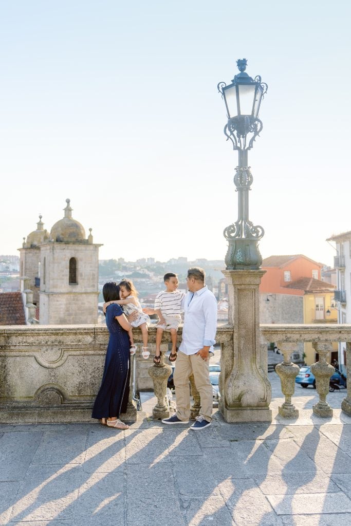 A family of four stands near a stone railing with an ornate lamppost, overlooking a scenic town with historic buildings in the background on a sunny day.