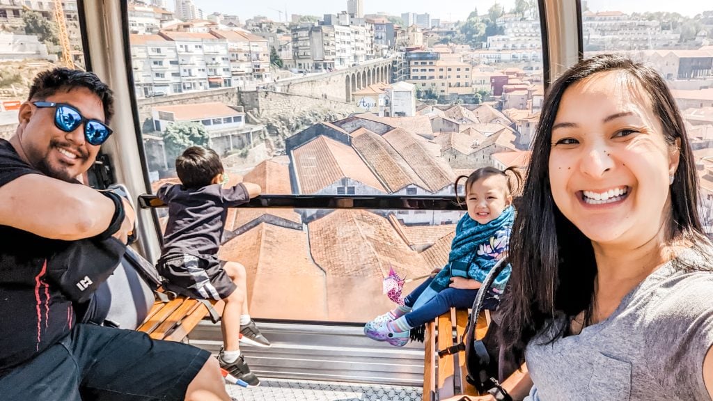A family of four rides in a Gaia cable car overlooking the city with red-tiled rooftops. The parents smile at the camera while the two children gaze out the window, marveling at the panoramic view.