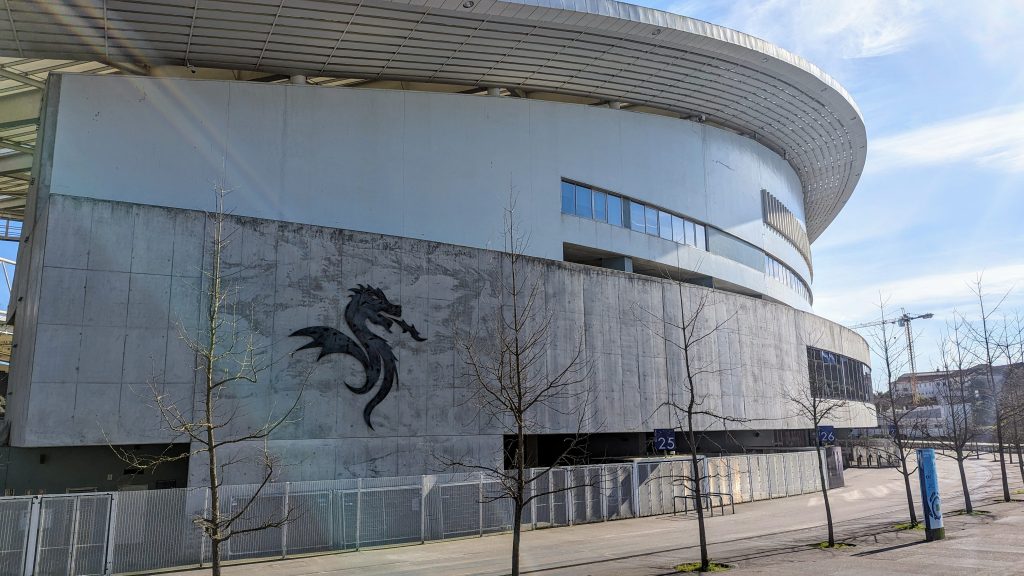 A large, curved stadium exterior with a dragon graphic on the concrete wall and bare trees in the foreground.