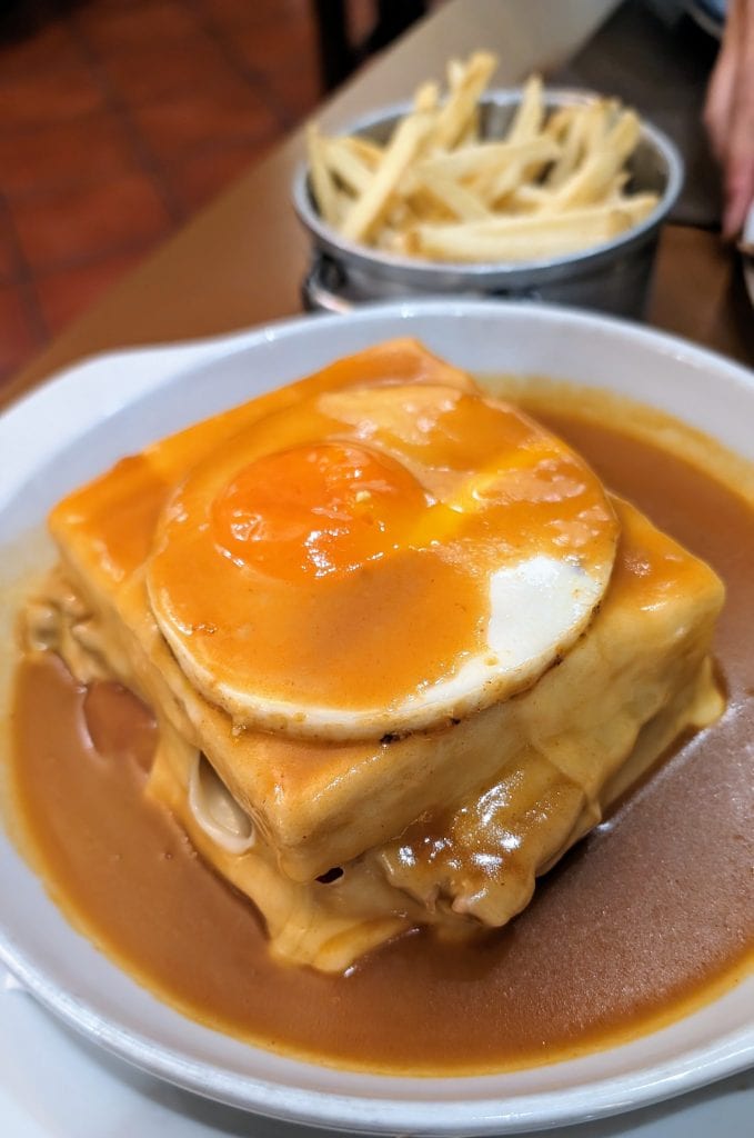 A dish of francesinha consisting of a sandwich covered in melted cheese and brown sauce, topped with a fried egg, with a side of French fries in the background.