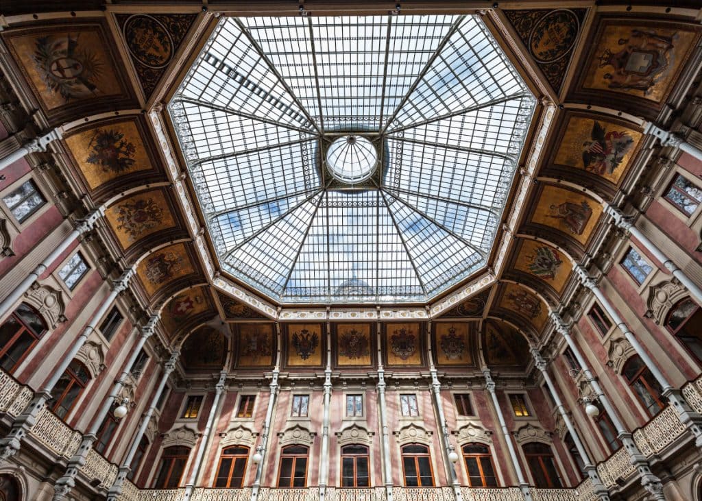 An ornate, multi-story interior with a large glass ceiling, decorated with detailed paintings and intricate architectural details. The ceiling features geometric patterns and a central circular window.