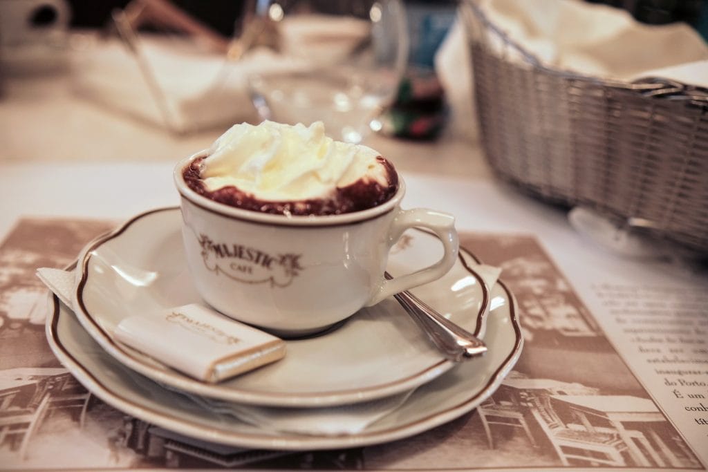 A cup of hot chocolate with whipped cream on top, served in a white cup and saucer, accompanied by a small packet of sugar and a spoon, on a table with a basket and other items in the background.