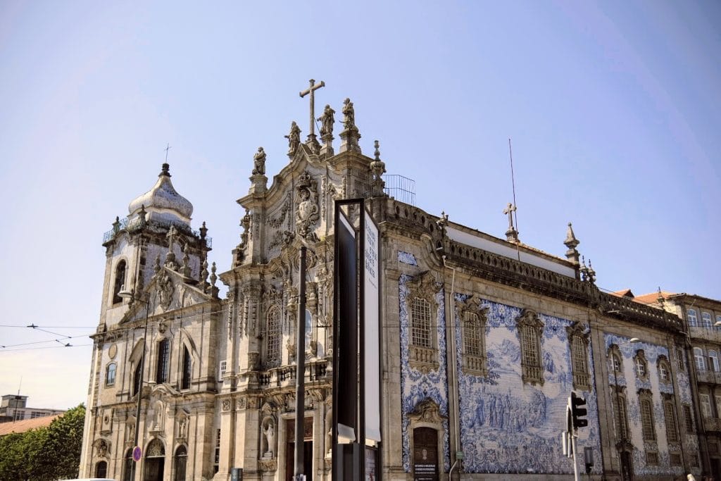 A historic baroque church, the Igreja do Carmo, boasts a detailed blue and white tiled facade under a clear sky on a sunny day. Visible in front are traffic lights and overhead tram cables.