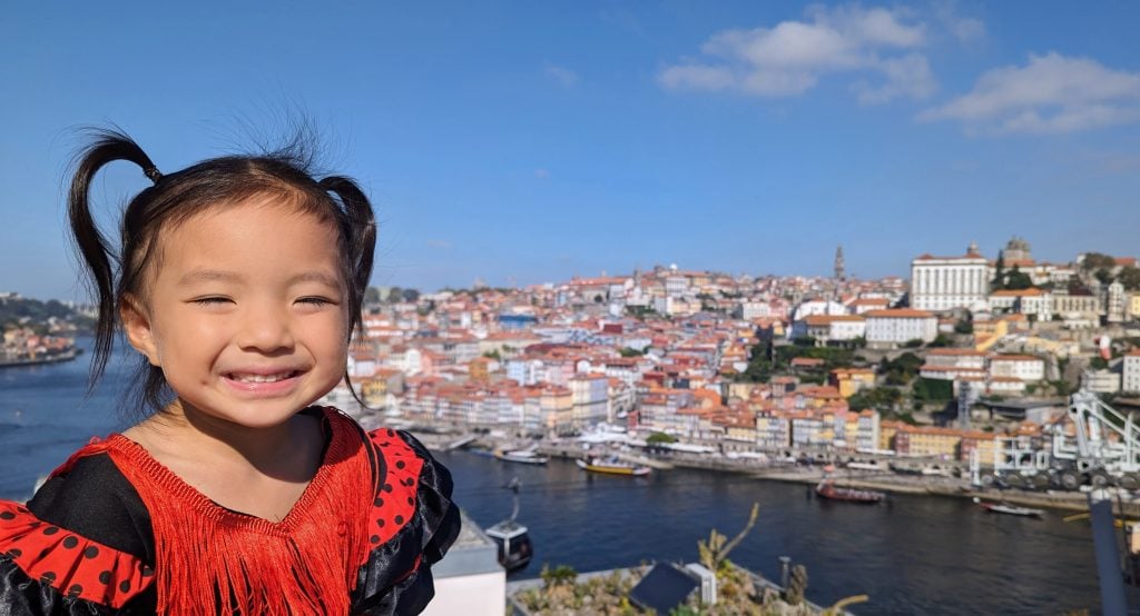 A young girl in a red and black dress smiles with the cityscape of Porto colorful buildings and the Douro river in the background under a clear blue sky.
