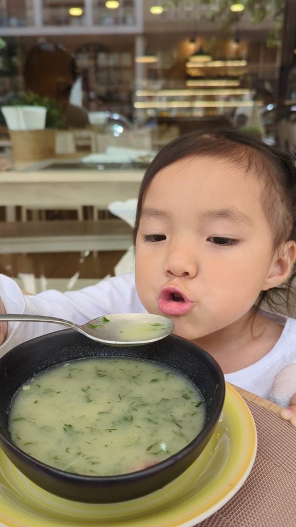 A young child in a white shirt is blowing on a spoonful of soup in front of a bowl of green soup at an indoor café.