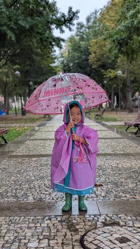 A child in a purple raincoat and green boots stands on a stone path holding a pink Minnie Mouse umbrella with trees and benches in the background.