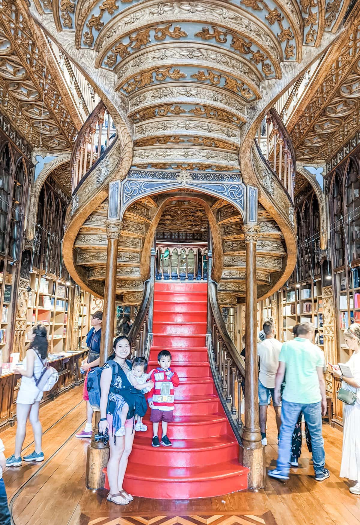 A woman and two children stand in front of an ornate spiral staircase with red steps inside a bookstore. Other people are in the background.