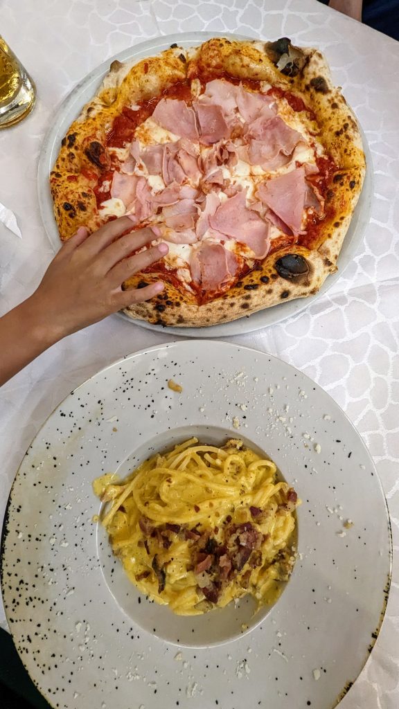 A hand reaches for a pizza topped with ham, placed on a table covered with a white tablecloth at Italy Caffe Gaia. Below the pizza is a plate of pasta carbonara, garnished with bacon bits and grated cheese.