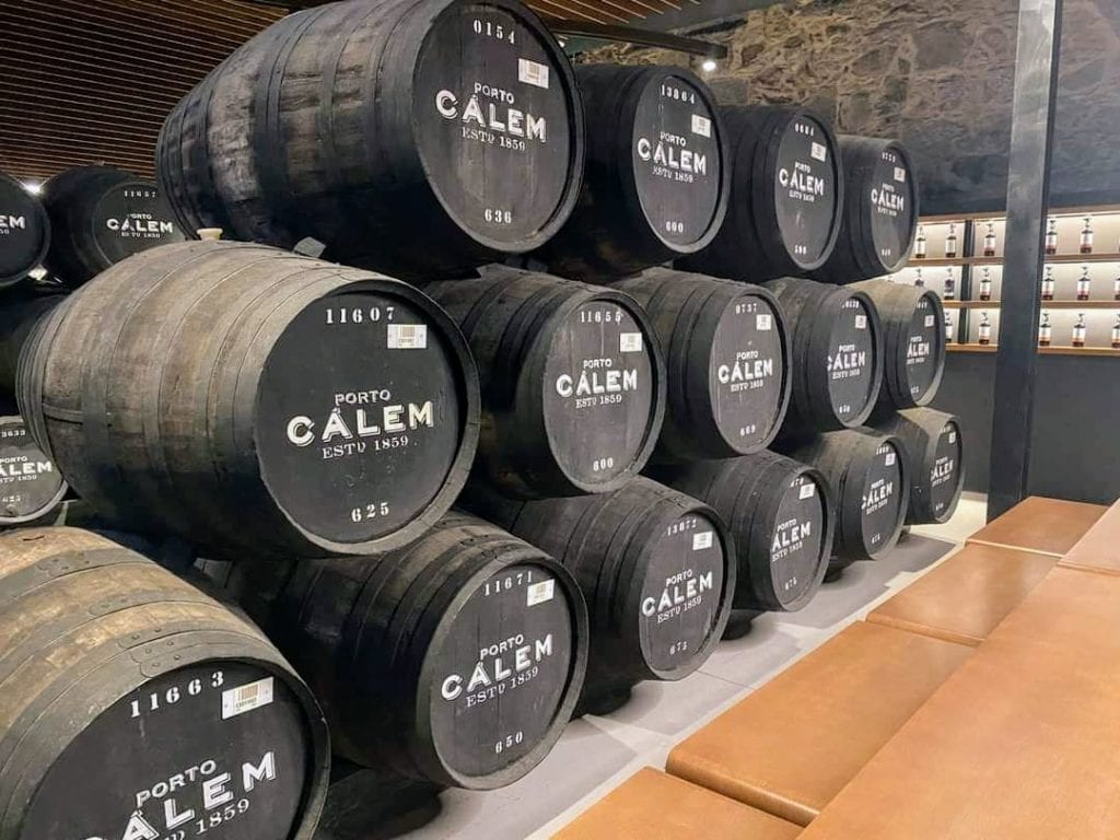 A stacked arrangement of large wooden barrels labelled "Porto Calem" in a storage area with seating.
