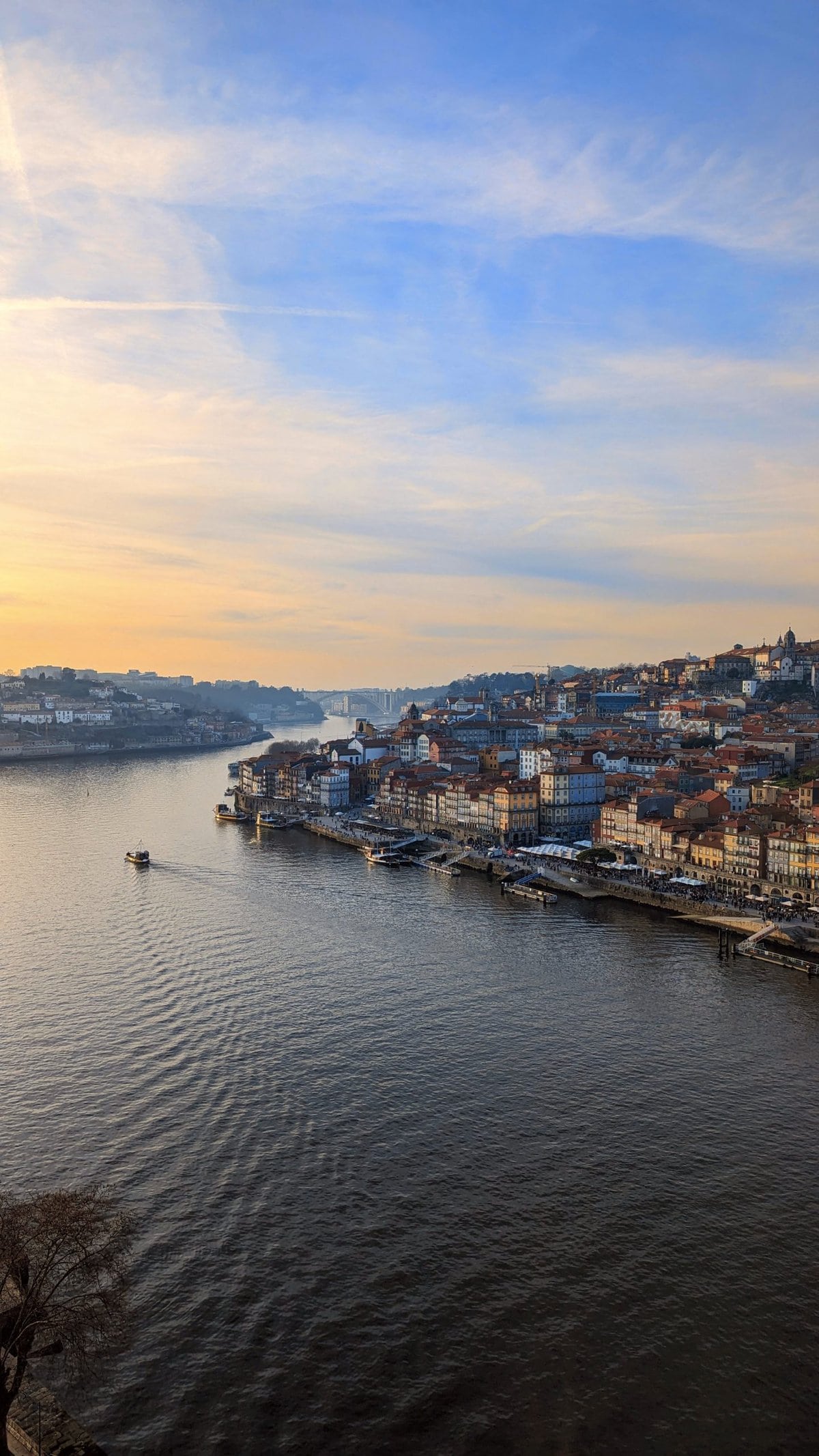 View of Porto from Dom Luis I Bridge with densely packed buildings along the shoreline, a boat sailing on the calm waters of Douro River, and a clear sky with scattered clouds at sunset.