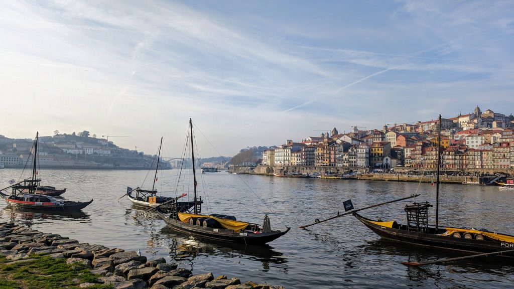 Traditional rabelo boats float on the calm river, with a scenic town featuring colorful buildings on a hillside in the background, under a partly cloudy sky. Stones line the riverbank in the foreground.