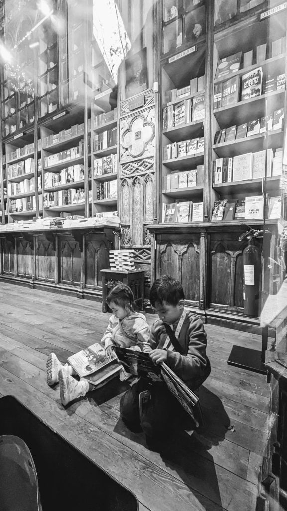 Two young children sitting on the floor of a bookstore, engrossed in reading. The background features tall bookshelves filled with various books. Black and white image.