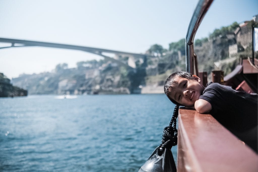 A smiling child leans over the railing of a boat on the Douro River, with a bridge and distant, blurred hills in the background.