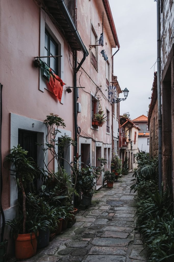 A narrow cobblestone alley in Porto's Ribeira district, lined with potted plants and residential buildings. Clothes hang from windows and balconies on both sides of the alley.