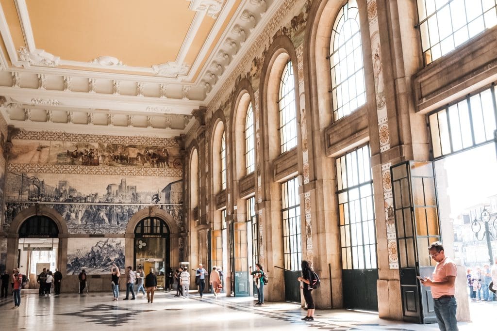 People standing and walking inside a large train station with high ceilings, arched windows, and decorative azulejo tiles on the walls.