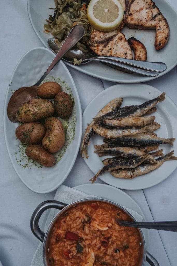 A meal consisting of a pot of seafood rice, a plate of grilled sardines, a dish of seasoned potatoes, and a platter of grilled swordfish with a lemon slice and greens.