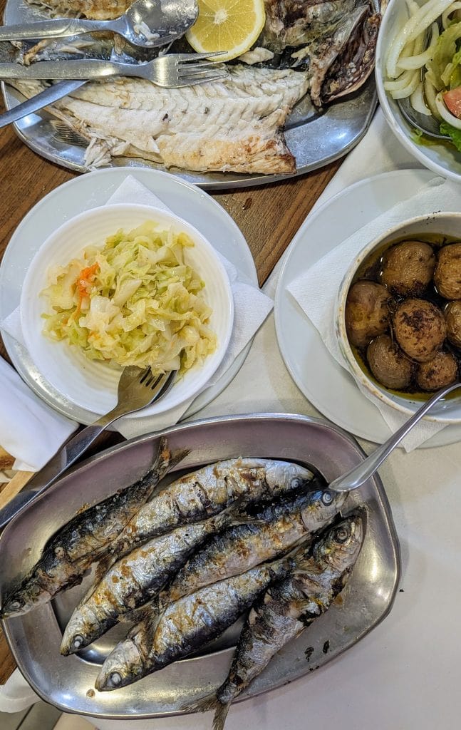 A table with various dishes including grilled sardines, boiled potatoes, a plate of vegetables.