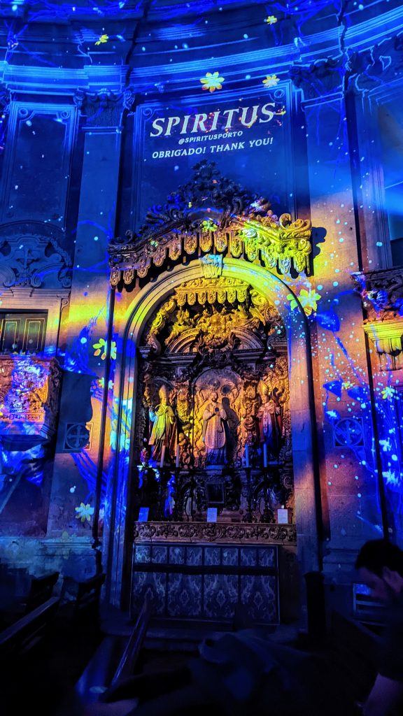 A church altar illuminated with colorful blue, yellow, and green lights displaying the word "SPIRITUS" and "OBRIGADO! THANK YOU!" on the wall above intricate religious statues.