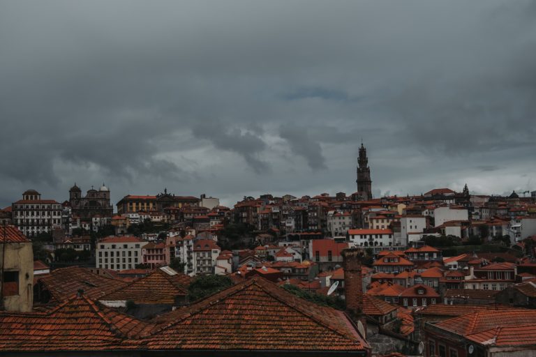 A cityscape with red-tiled rooftops under a cloudy sky, highlighting a prominent tower among closely clustered buildings.