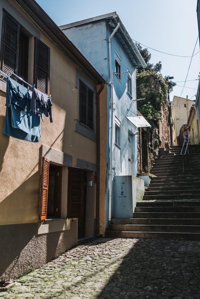 Narrow cobblestone alley in the Ribeira district with two houses on either side; laundry hangs from a line on the left, and a steep staircase ascends on the right. Sunlight casts shadows on the buildings.