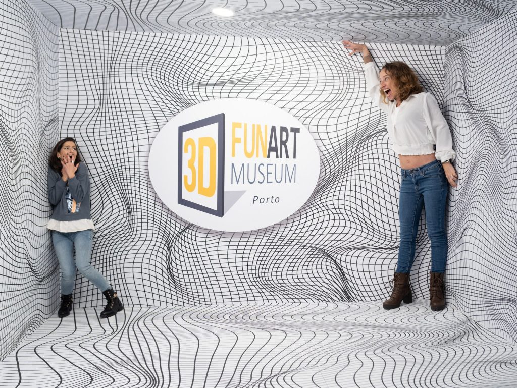 Two women pose playfully in a 3D illusion room with dynamic black-and-white grid patterns at the 3D Fun Art Museum in Porto. One points to the logo, and the other covers her mouth in surprise.