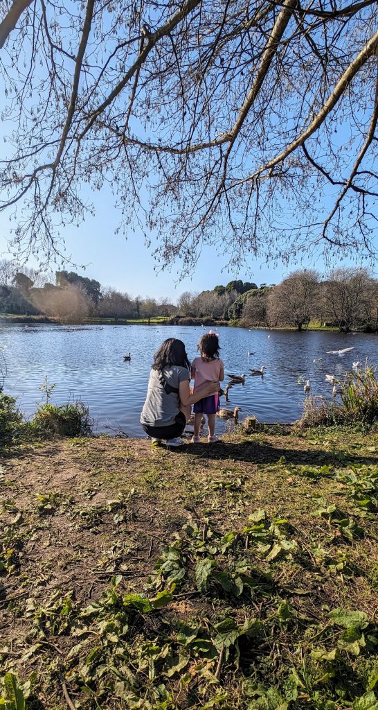 An adult and a child kneel by a lakeshore watching ducks swim on a calm, sunny day, with trees and foliage partially covering the view.