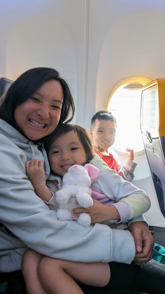 A woman with a child on her lap holding a stuffed animal, sitting on an airplane. Another child is seated beside them near the window.