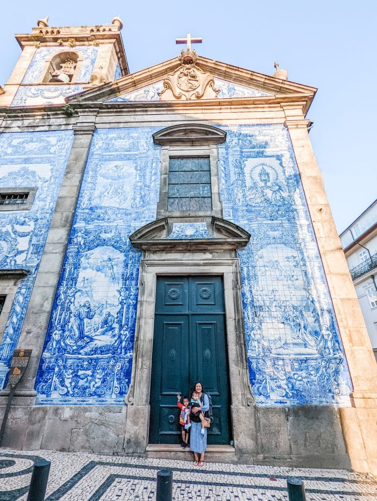 Three people stand in front of a historic building with blue and white tilework façade, featuring detailed artwork. The building has a green double door and a cross atop its peaked roof.