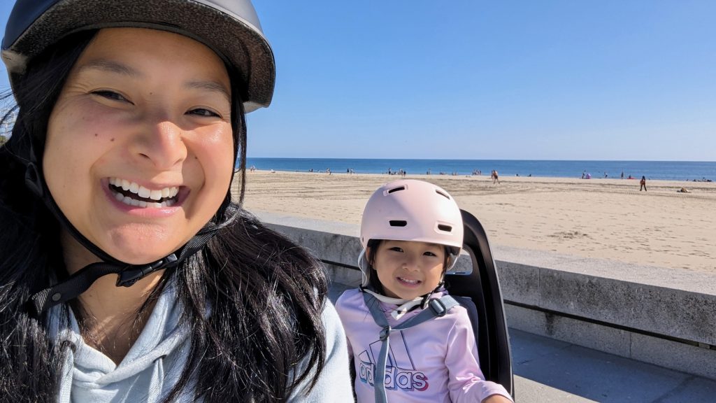 Two people wearing helmets smile while sitting near a sandy beach with the ocean in the background on a sunny day.