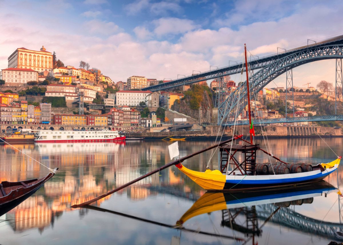 A vibrant coastal cityscape in Porto, Portugal showcases traditional boats on a calm river, with historic buildings and the Dom Luís I Bridge in the background under a partly cloudy sky.