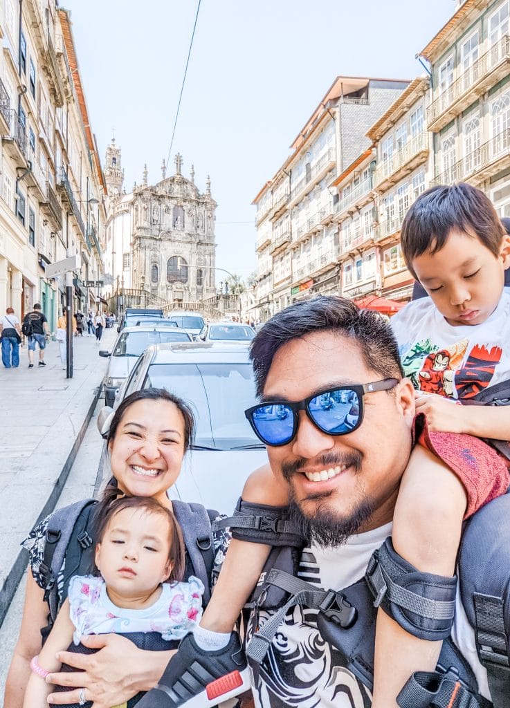 A smiling family with two children, one being carried in a front backpack and another on an adult's shoulders, poses on a street with old buildings and a church in the background.